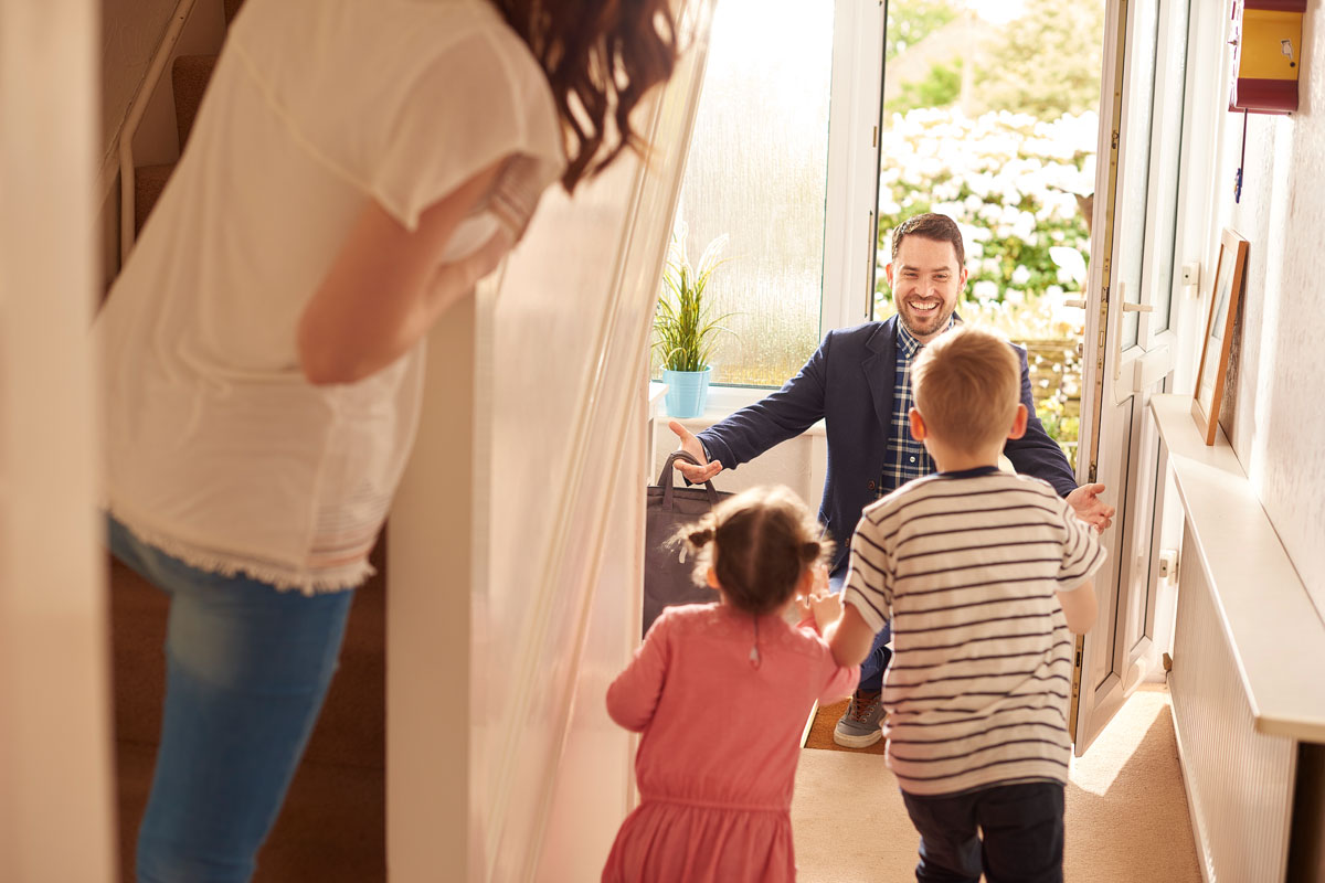 A smiling man in a suit greeting two young children at the front doorway of a home